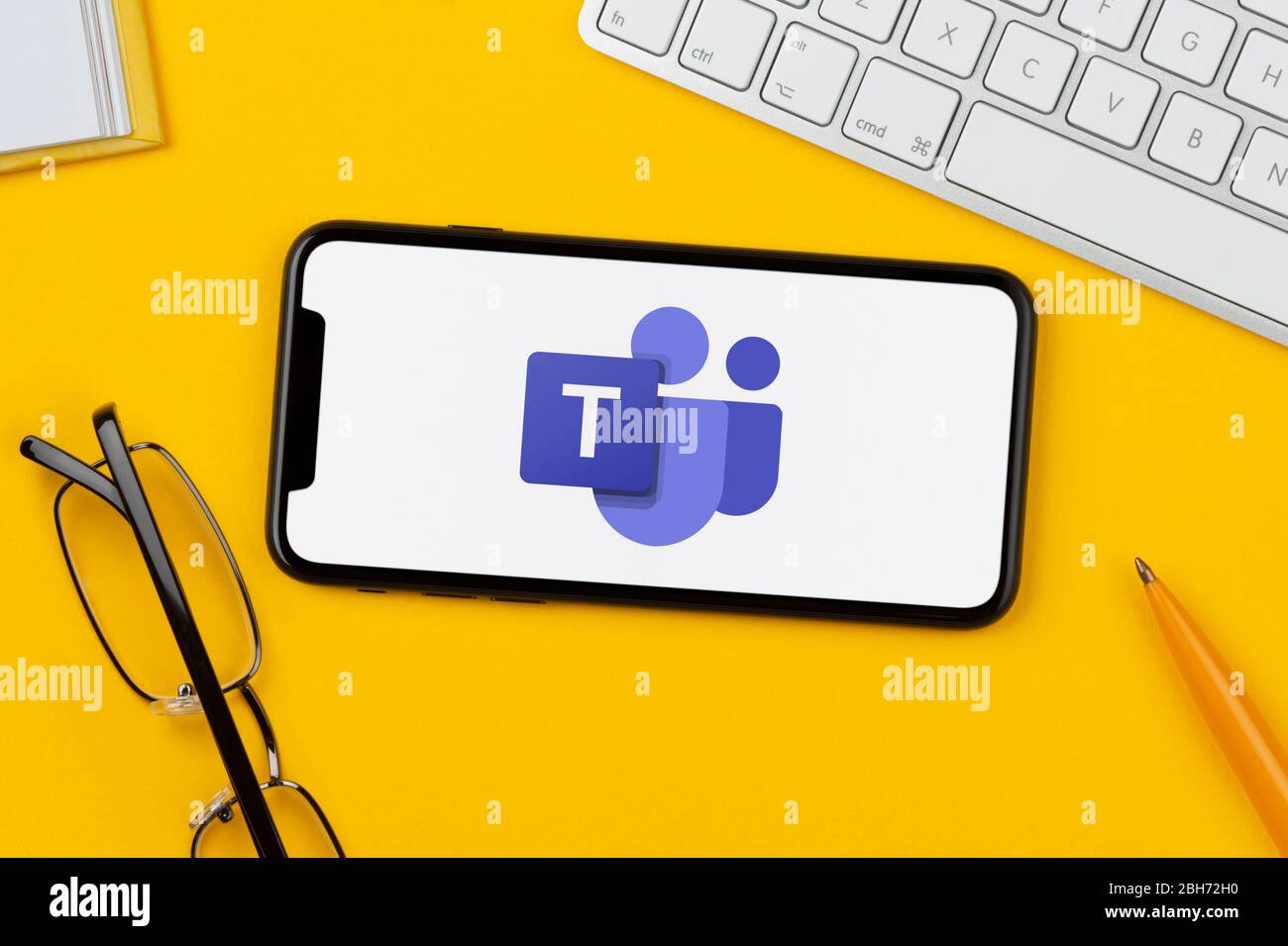 Ein Smartphone mit dem Microsoft Teams Logo liegt auf einem gelben Hintergrund zusammen mit Tastatur, Brille, Stift und Buch (nur für redaktionelle Zwecke). Stockfoto