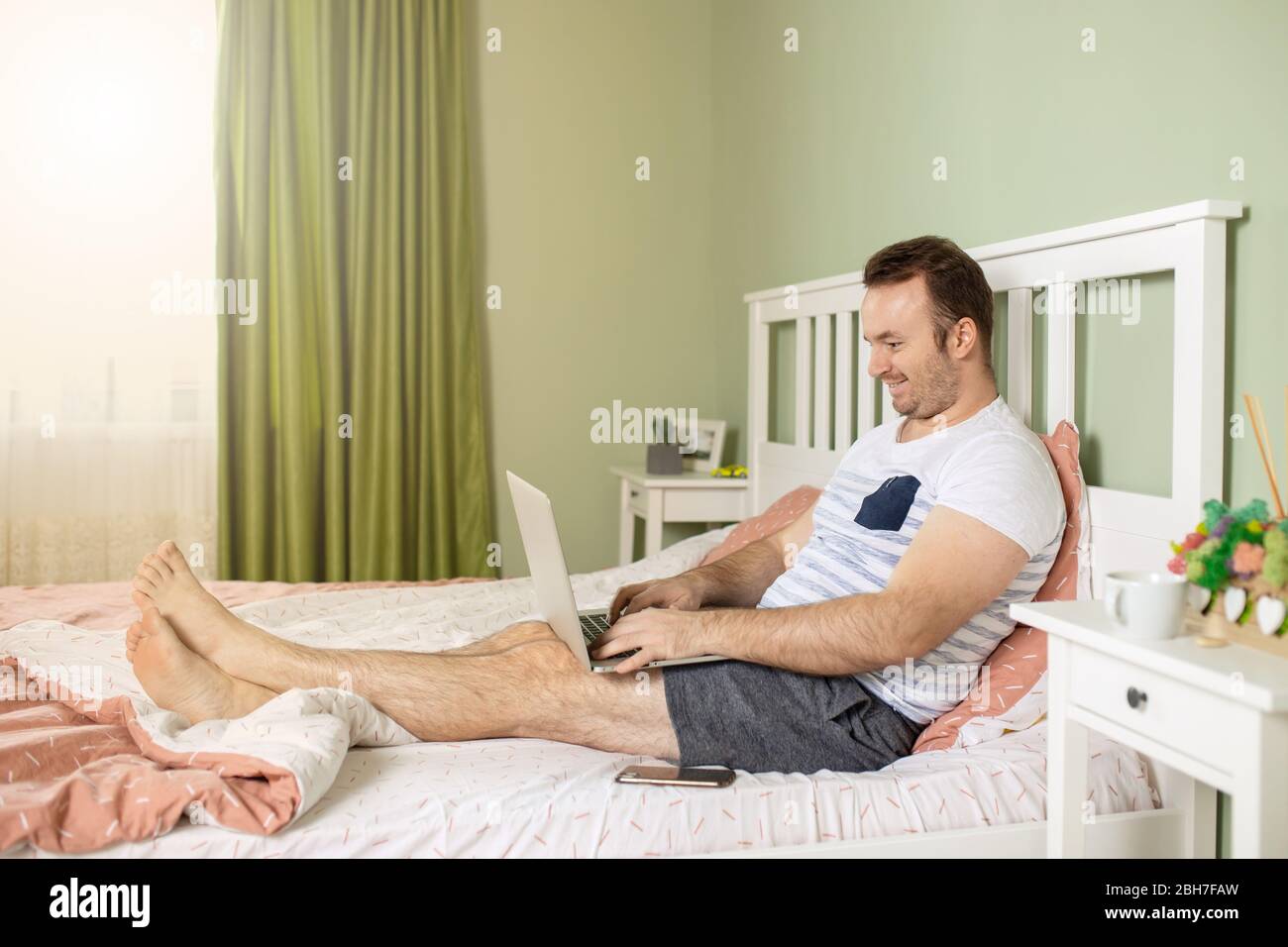 Der Mann mit einem Laptop im Bett erhielt schlechte Nachrichten, er ist verwirrt, hält die Hand am Kopf und beobachtet den Bildschirm in schockierten Augen. Stockfoto