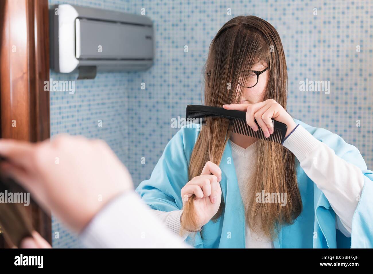 Ein schönes junges Mädchen hält einen Kamm, um ihr Haar während der Lockdown von Coronavirus schneiden, Blick in Spiegel. DIY Haarschnitt während der Epidemie o Stockfoto