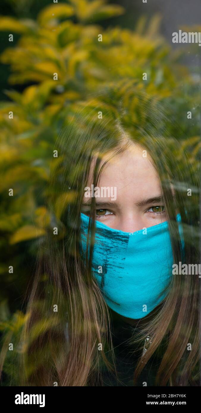 Teenager-Mädchen hinter Fenster tragen Hause gemacht Gesichtsmaske. Bleiben Sie zu Hause einsam. Stockfoto