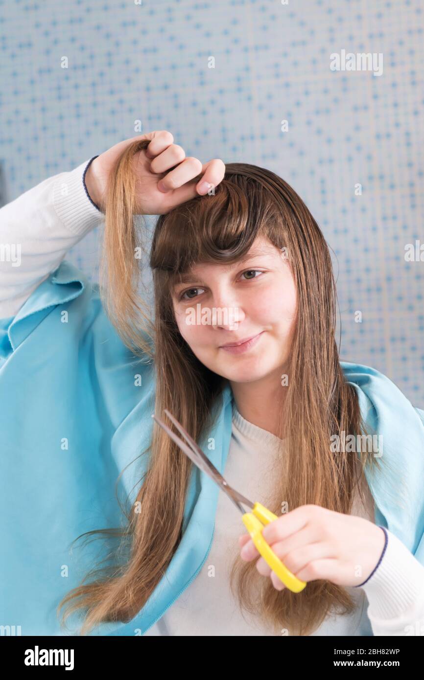 Ein schönes junges Mädchen hält eine Schere, um sich während des Lockdown  von Coronavirus die Haare zu schneiden und im Spiegel zu schauen. DIY Haare  schneiden während der Epidemii Stockfotografie - Alamy