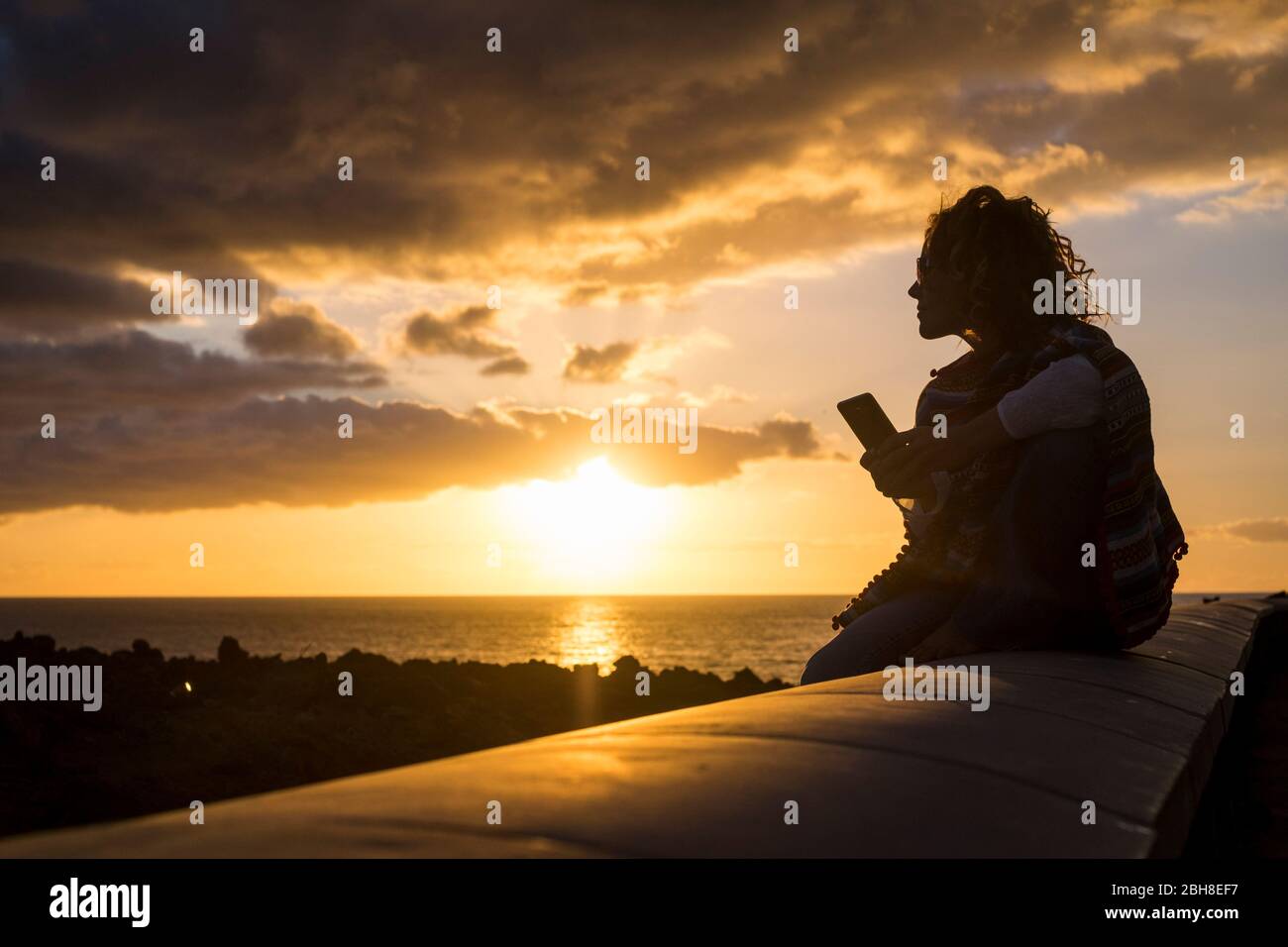Schöne Frau in Silhouette während eines goldenen erstaunlichen Sonnenuntergang auf dem Meer mit einem technolofy Handy sitzen auf einem langen Stein Beanch. Schöne Freizeit im Urlaub schreiben Freunde Stockfoto