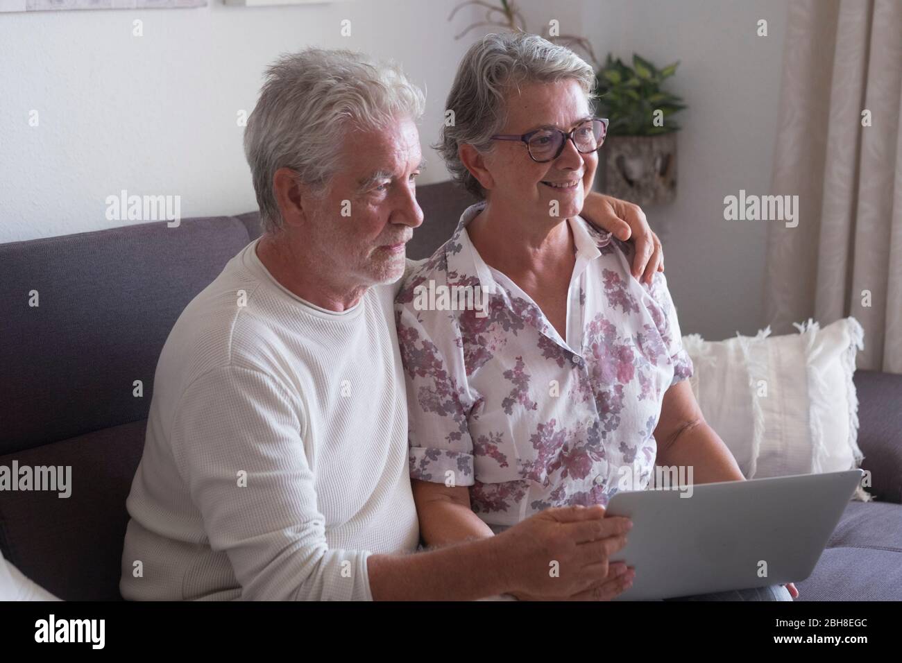 Ältere Paare zu Hause surfen auf Laptop-Computer - Frau lächelnd und Mann ernst sowohl auf der Suche nach etwas im Internet und verwenden Sie ein Notebook setzen Sie sich auf das Sofa Stockfoto