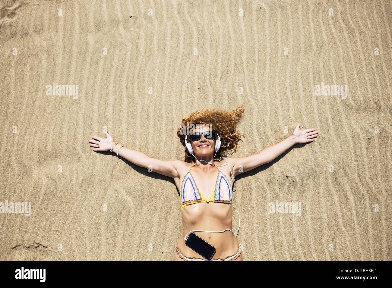 Schöne mittlere Alter Tourist Frau genießen den warmen Sand und die Sommertage des Urlaubs legen und Musik mit Handy-Technologie und Telefon hören. Urlaub und Glück Bild Stockfoto