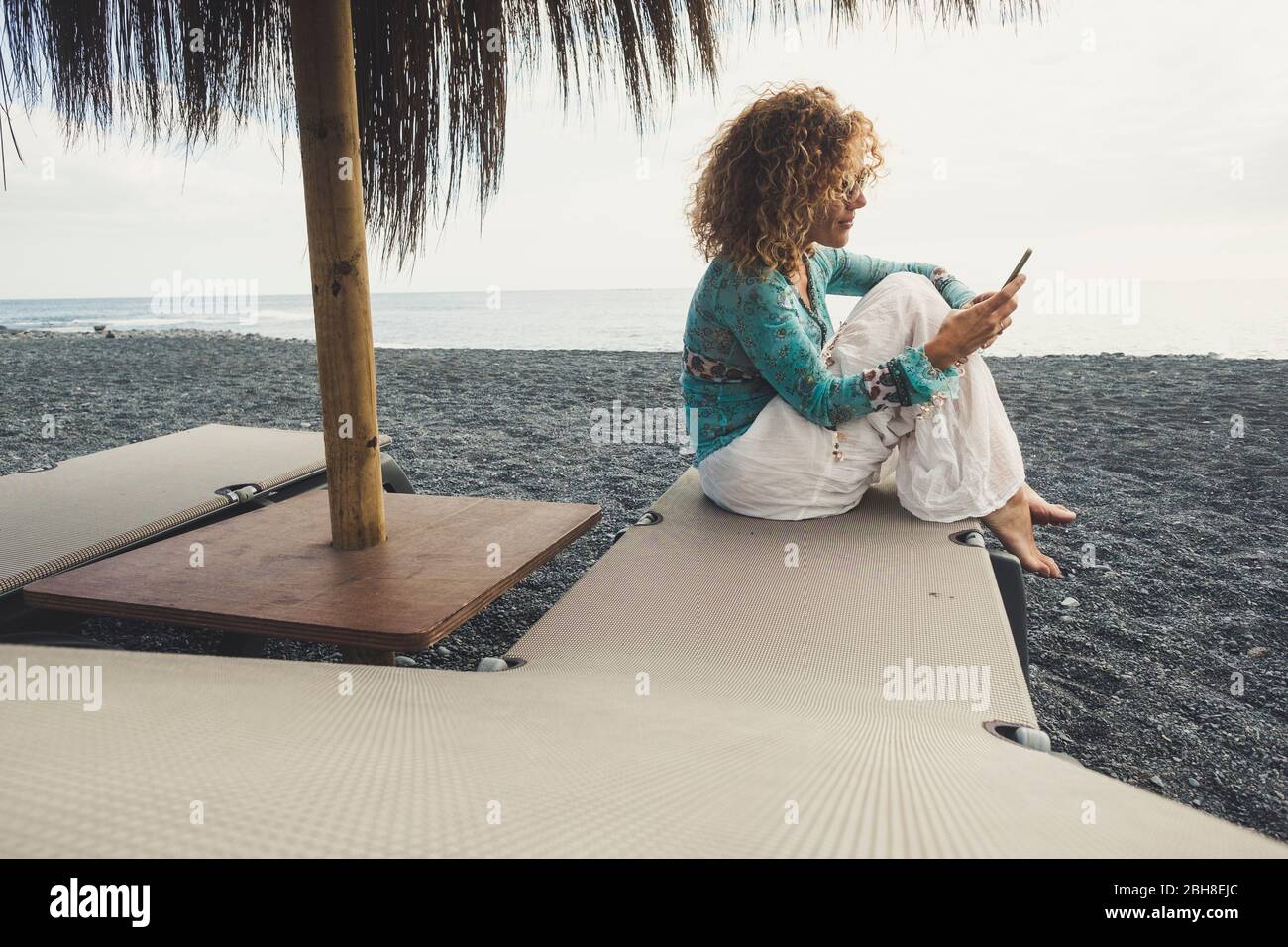 Eine nette Frau am Strand setzte sich auf die Sitze mit Meer und Sand um sie herum und überprüfte das Telefon Stockfoto