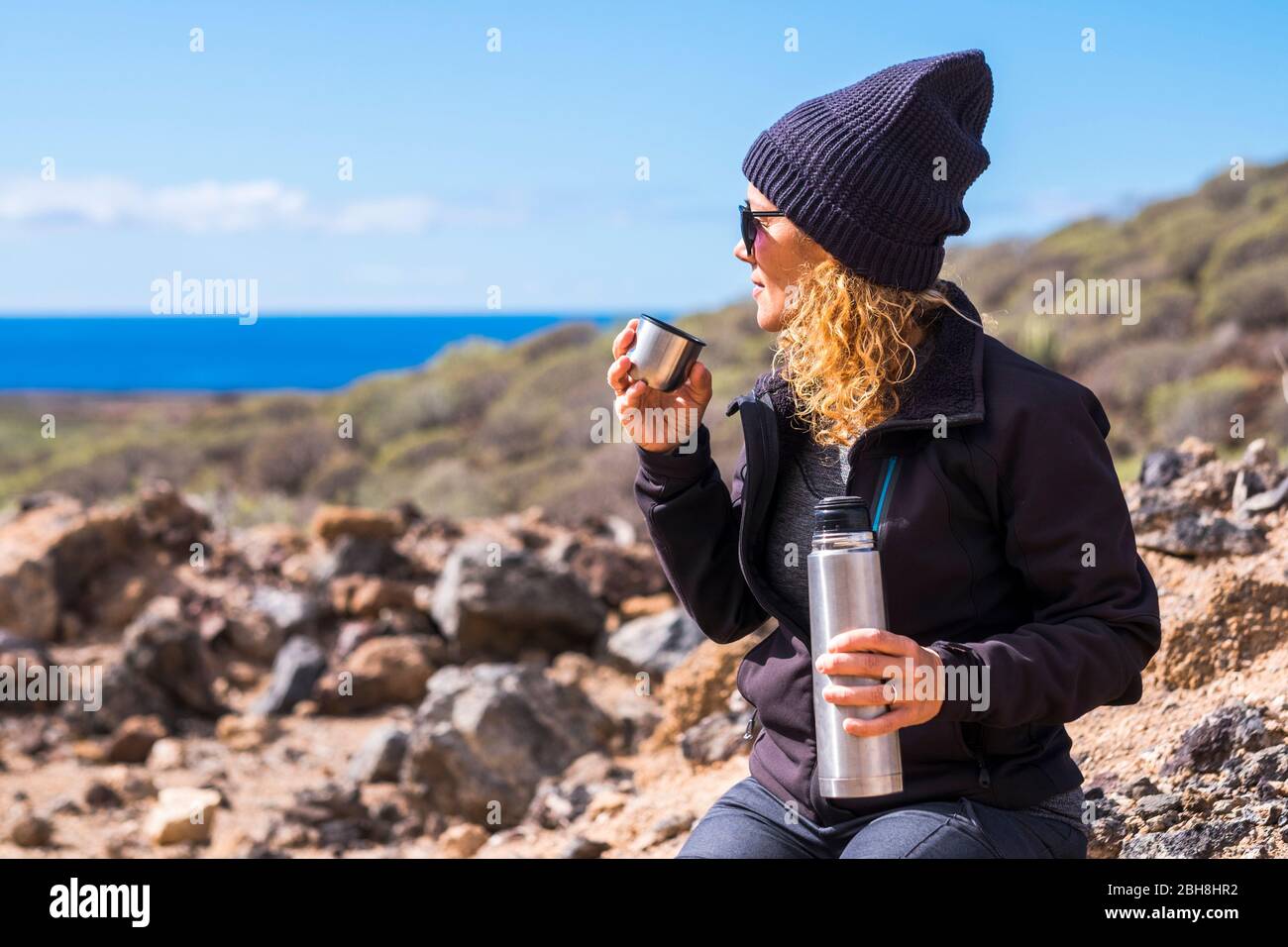 Hübsche Frau mit blonden lockigen Haaren und schwarzem warmen Hut genießen die Outdoor-Aktivität trinken etwas Tee oder Kaffee und Blick auf das Meer im Hintergrund - himer und Trekking-Konzept für freie Menschen Stockfoto