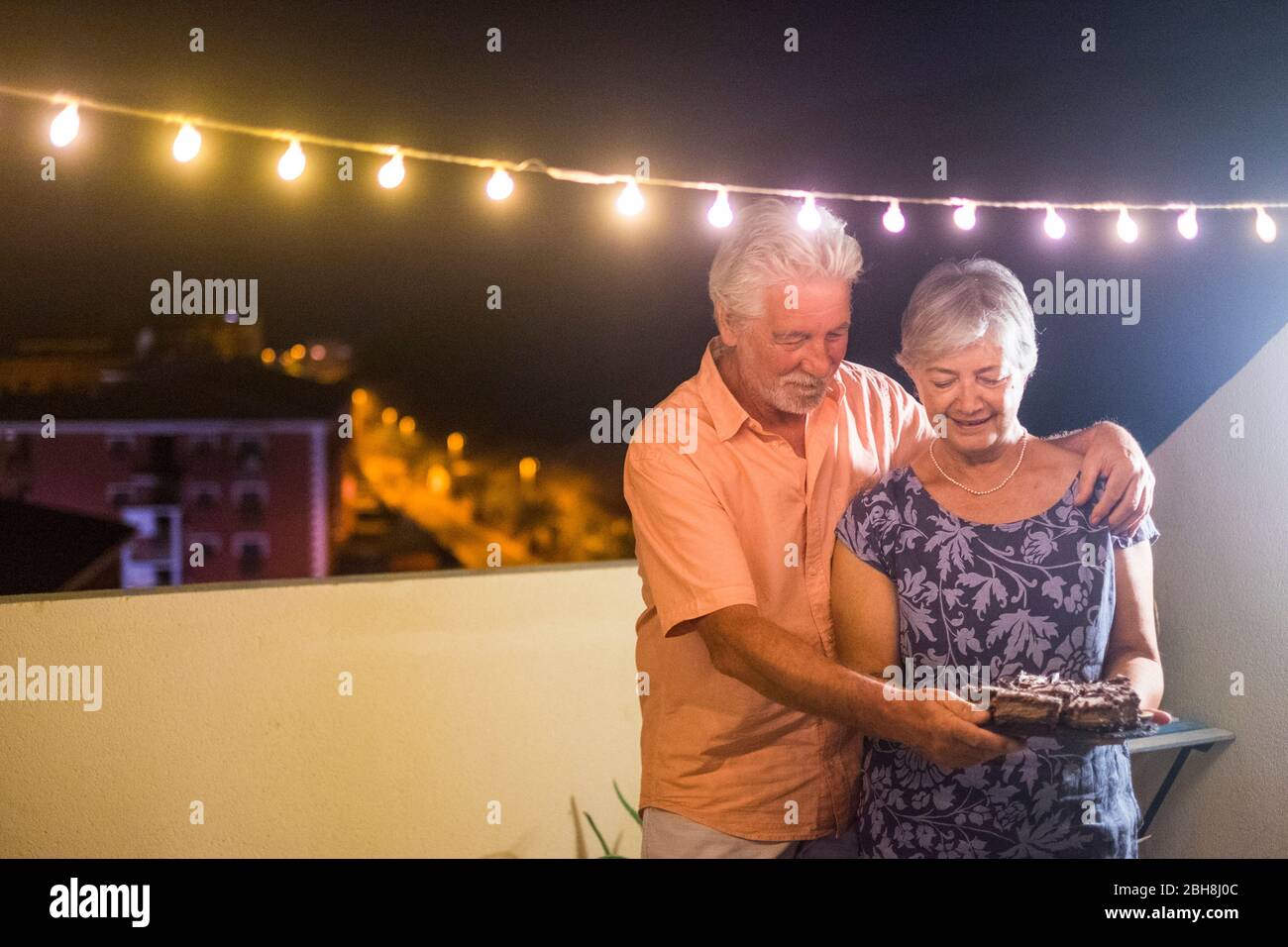 Happy Senior Erwachsene Paar mit einem Schokoladenkuchen nach dem Abendessen zu Hause im Freien auf der Terrasse mit Party Glühbirne Licht - Geburtstagskonzept oder Neujahr Feier für Erwachsene Menschen - Stockfoto