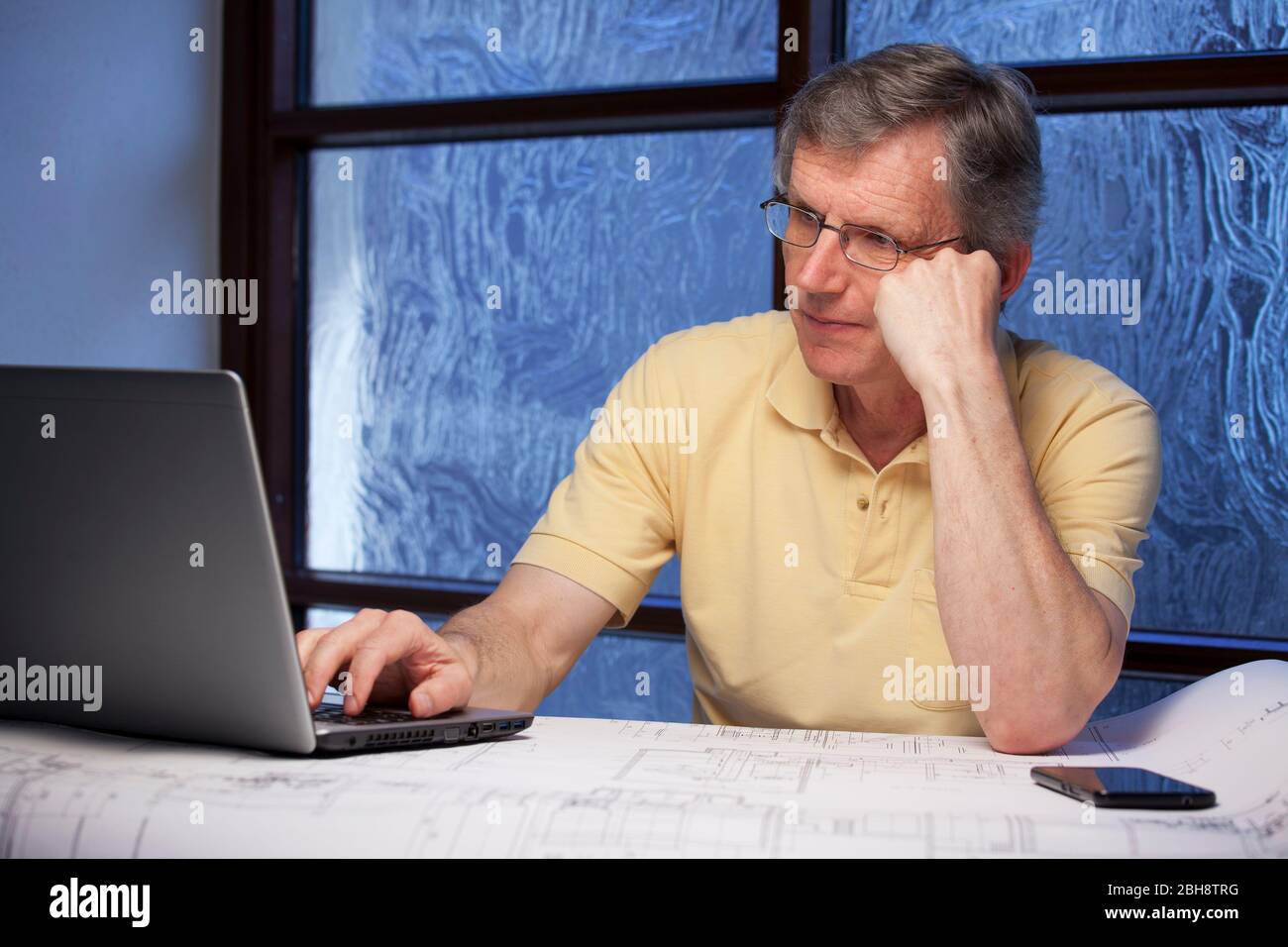 Leitender nachdenklicher Geschäftsmann oder Ingenieur, der von zu Hause aus an einem Schreibtisch mit Laptop, Plan und Handy arbeitet - konzentrieren Sie sich auf das Gesicht Stockfoto