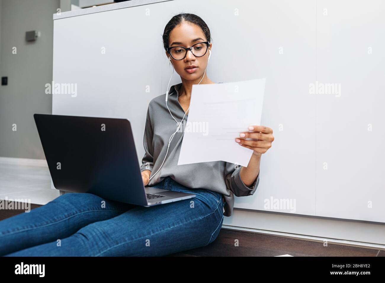 Junge Frau auf dem Boden an der Küchentheke mit Dokument in der Hand und mit Laptop Stockfoto