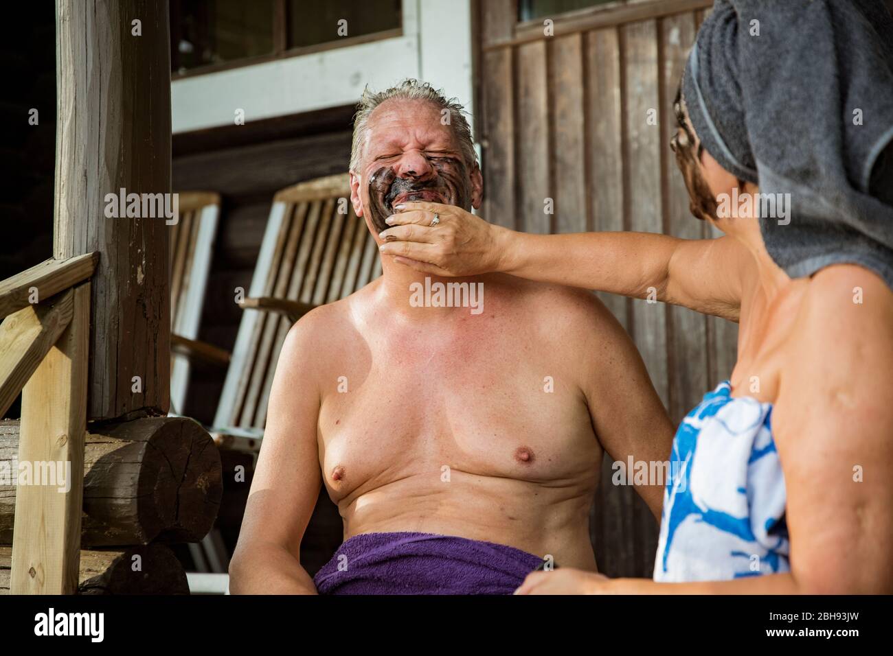 Gealtertes Paar mit schwarzer Gesichtsmaske nach finnischer Sauna. Reifer Mann und Frau in Handtüchern sitzen auf Holztreppen, entspannen, lachen und Spaß haben. Stockfoto