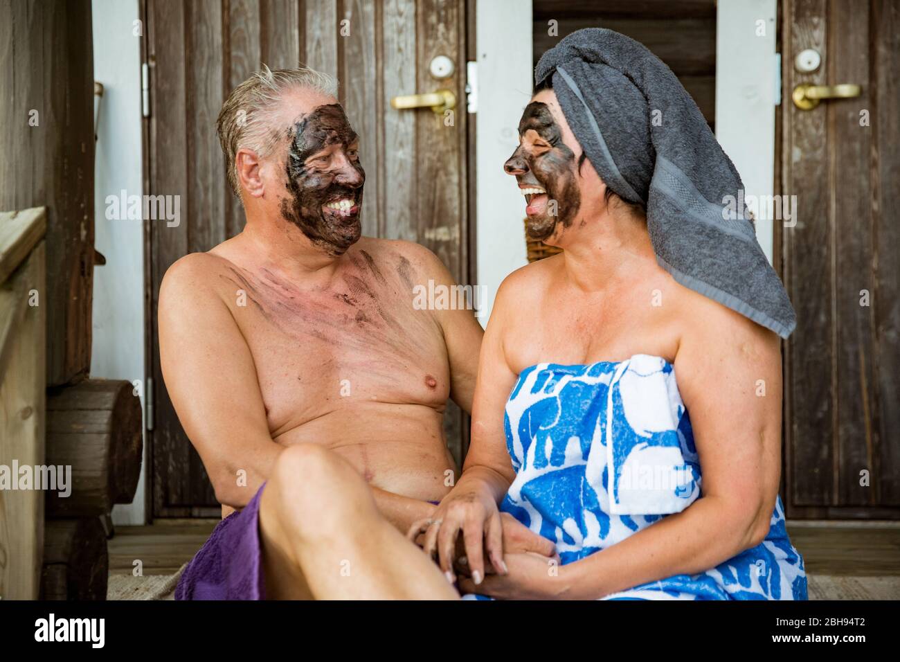 Gealtertes Paar mit schwarzer Gesichtsmaske nach finnischer Sauna. Reifer Mann und Frau in Handtüchern sitzen auf Holztreppen, entspannen, lachen und Spaß haben. Stockfoto