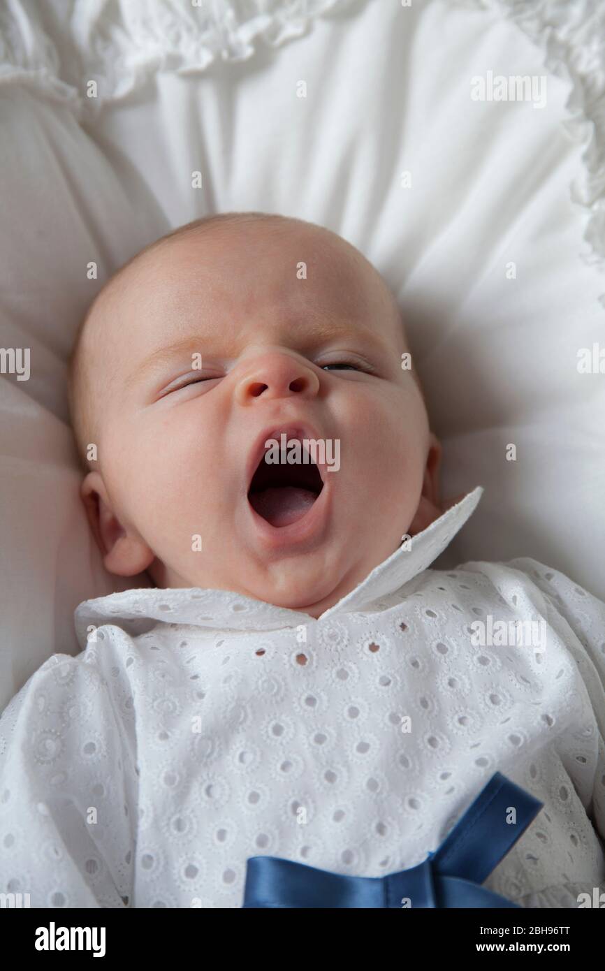 Zwei Monate altes Baby mit Taufkleid gähnt, auf weißem Kissen liegend, Stockfoto