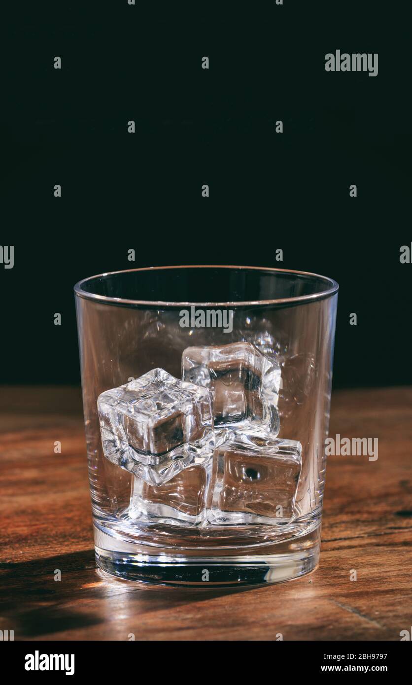 Kristall leeres Whiskyglas mit Eiswürfeln auf Holztheke. Vertikales Porträt eines klaren transparenten Glasgefäßbecher voller Eissteine auf dunkel Stockfoto
