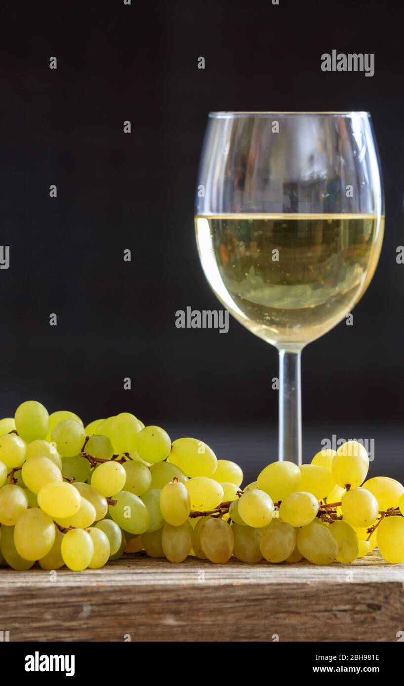 Weißweinglas neben einem Traubenstrauß auf einem Holztisch. Vertikales Porträt von Alkohol und Obst auf schwarzem Hintergrund. Stockfoto