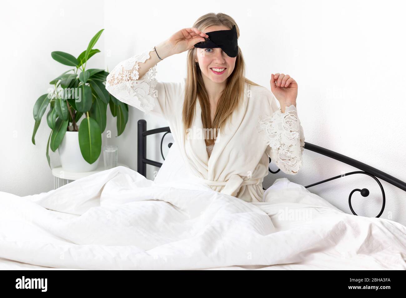 Eine Europäerin mit blonden Haaren sitzt auf einem weißen Bett, sie ist gerade aufgewacht. Sie ist in leichte Kleidung und eine Schlafmaske gekleidet. Fauler Wochenendmorgen Stockfoto