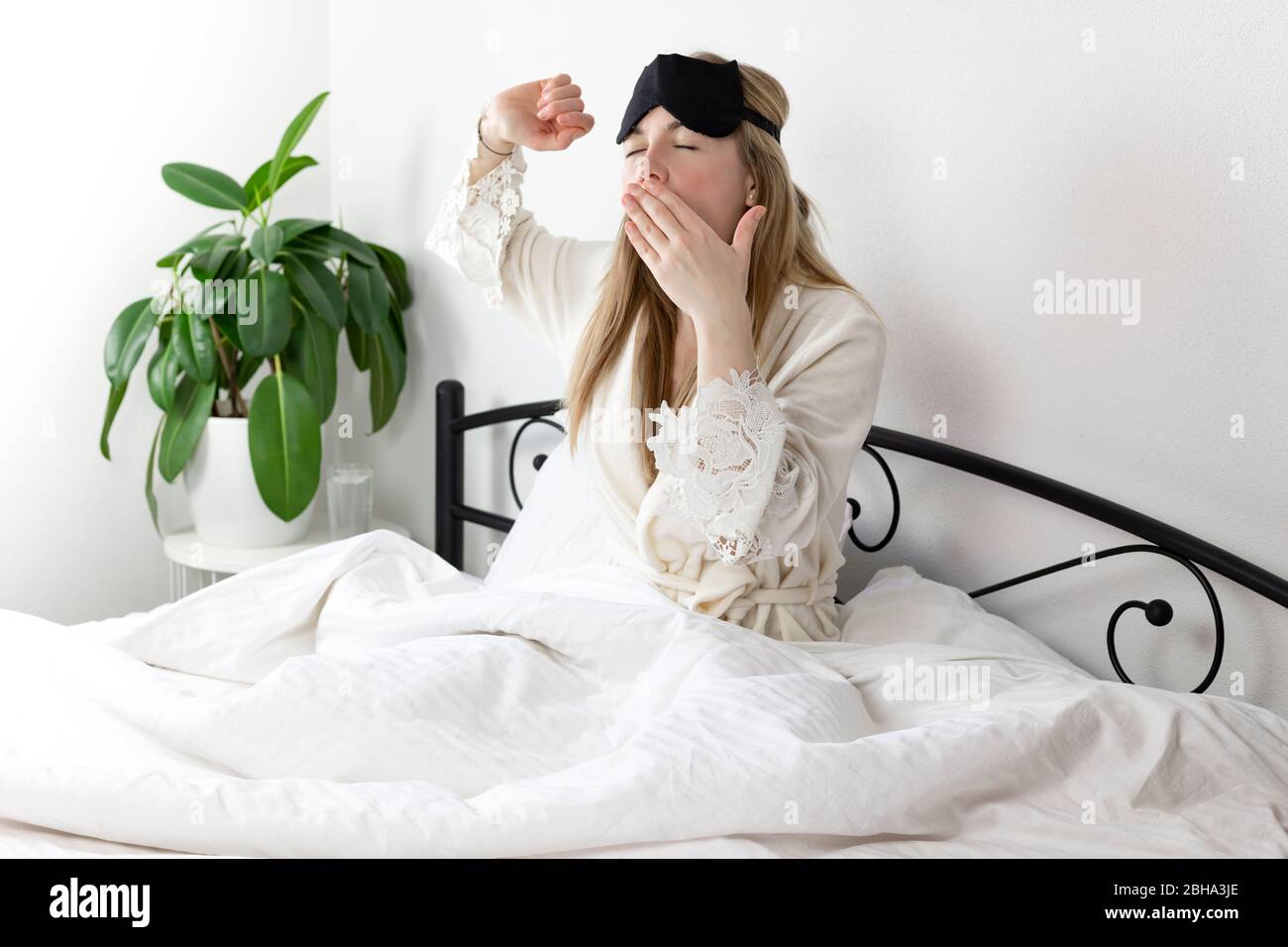 Eine Europäerin mit blonden Haaren sitzt auf einem weißen Bett, sie ist gerade am Morgen aufgewacht. Sie ist in leichte Kleidung und eine Schlafmaske gekleidet. Sie d Stockfoto