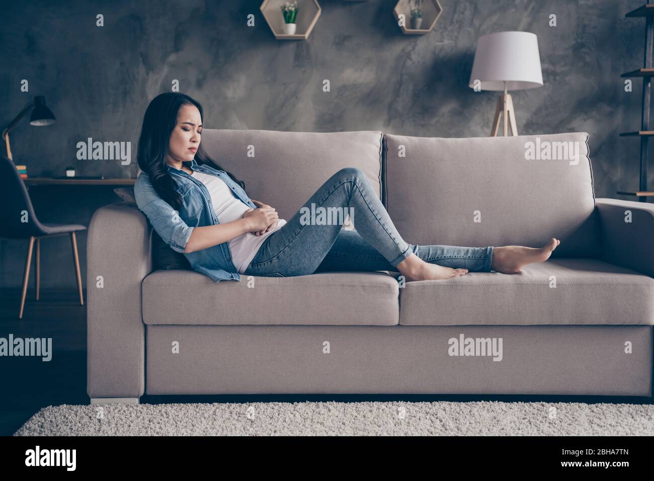 Profilfoto von häuslichen ziemlich unzufrieden asiatische Dame liegen bequeme Couch zu Hause bleiben Quarantäne Zeit halten Hände auf Bauch leiden Schmerzen Magen pms Stockfoto