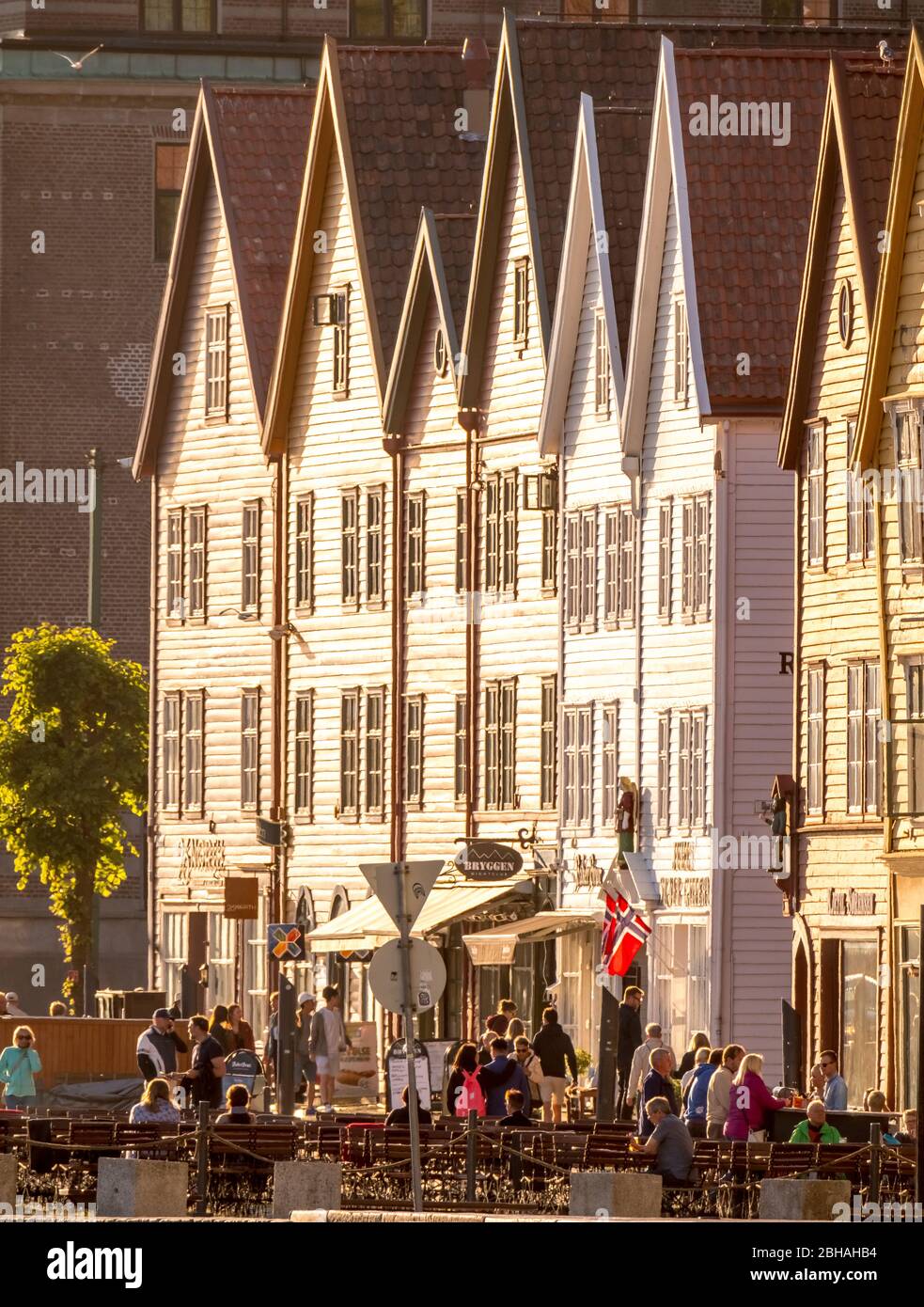 Touristen gehen, andere sitzen an Tischen auf Stühlen vor einem weißen Holzhaus mit Geschäften im Hafen von Bryggen, Bergen, Hordaland, Norwegen, Skandinavien, Europa Stockfoto