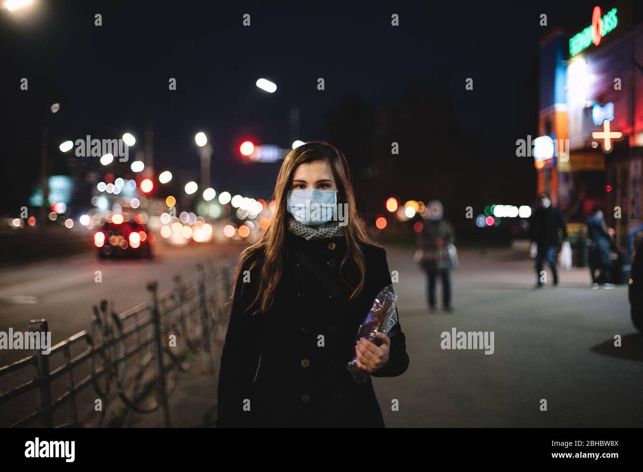 Porträt einer jungen Frau mit Gesicht medizinische Maske hält Brot, während auf der Straße der Stadt in der Nacht Stockfoto