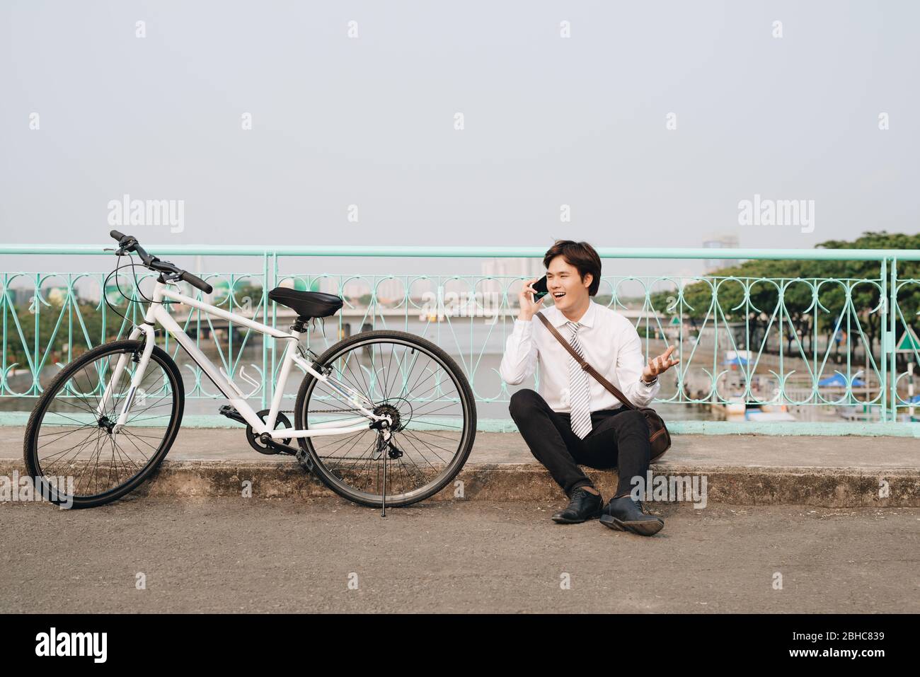 Jungen asiatischen Mann per Handy beim Sitzen auf dem Boden in der Nähe des Rad, im Freien. Stilvolle Kleidung tragen. Stockfoto