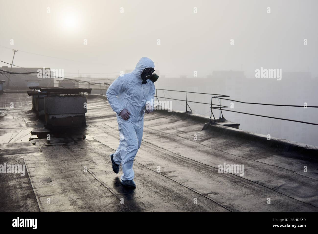 Mann trägt weiße Overall und Gasmaske, läuft auf dem Dach eines städtischen Gebäudes mit dem Rücken zu einer aufgehenden Sonne bei Nebel Wetter, Konzept der Umweltproblem Stockfoto
