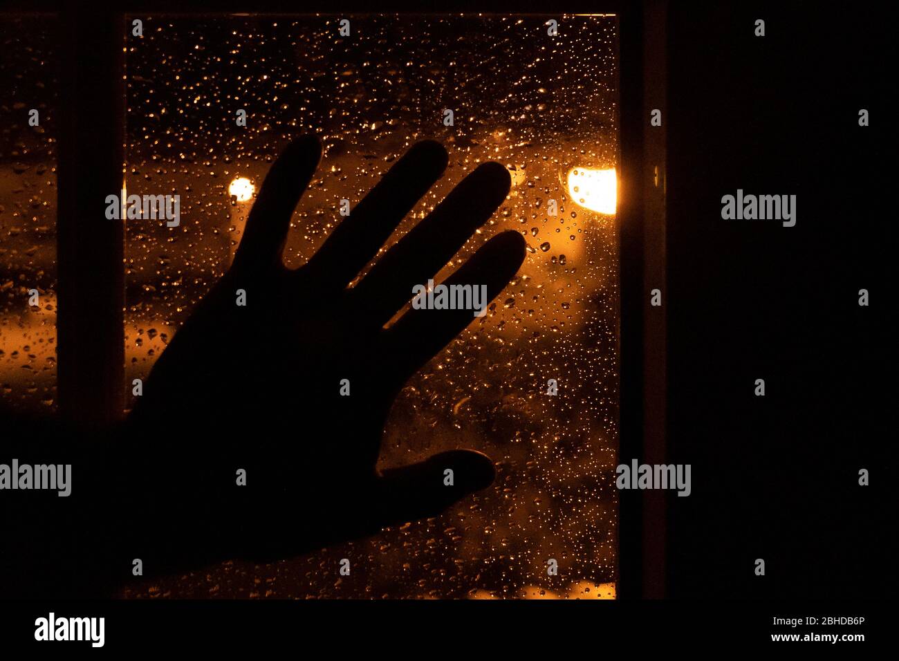 Silhouette einer Hand, die nachts am Fenster mit dem warmen Licht der Straßenlampen und mit Wassertropfen von einem regnerischen Tag ruht. Trauriges Bild von mel Stockfoto