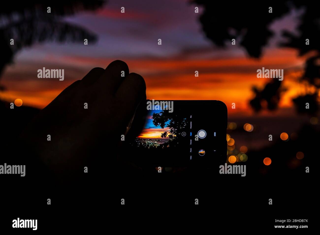 Ein Foto einer Hand, die ein Mobiltelefon hält, das ein Bild eines Sonnenuntergangs im Fokus auf dem Bildschirm im Querformat zeigt ein verschwommener Hintergrund Stockfoto
