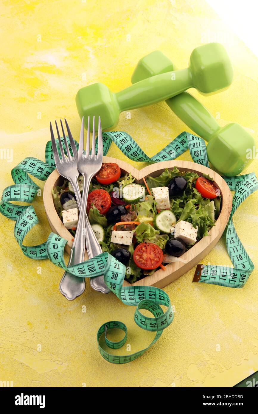 Authentischer frischer Salat in einer herzförmigen Tasse aus Holz mit Hanteln, Trainingsausrüstung, Messband auf dem Tisch. Gesunde Lebensführung, gutes Gesundheits-Concep Stockfoto