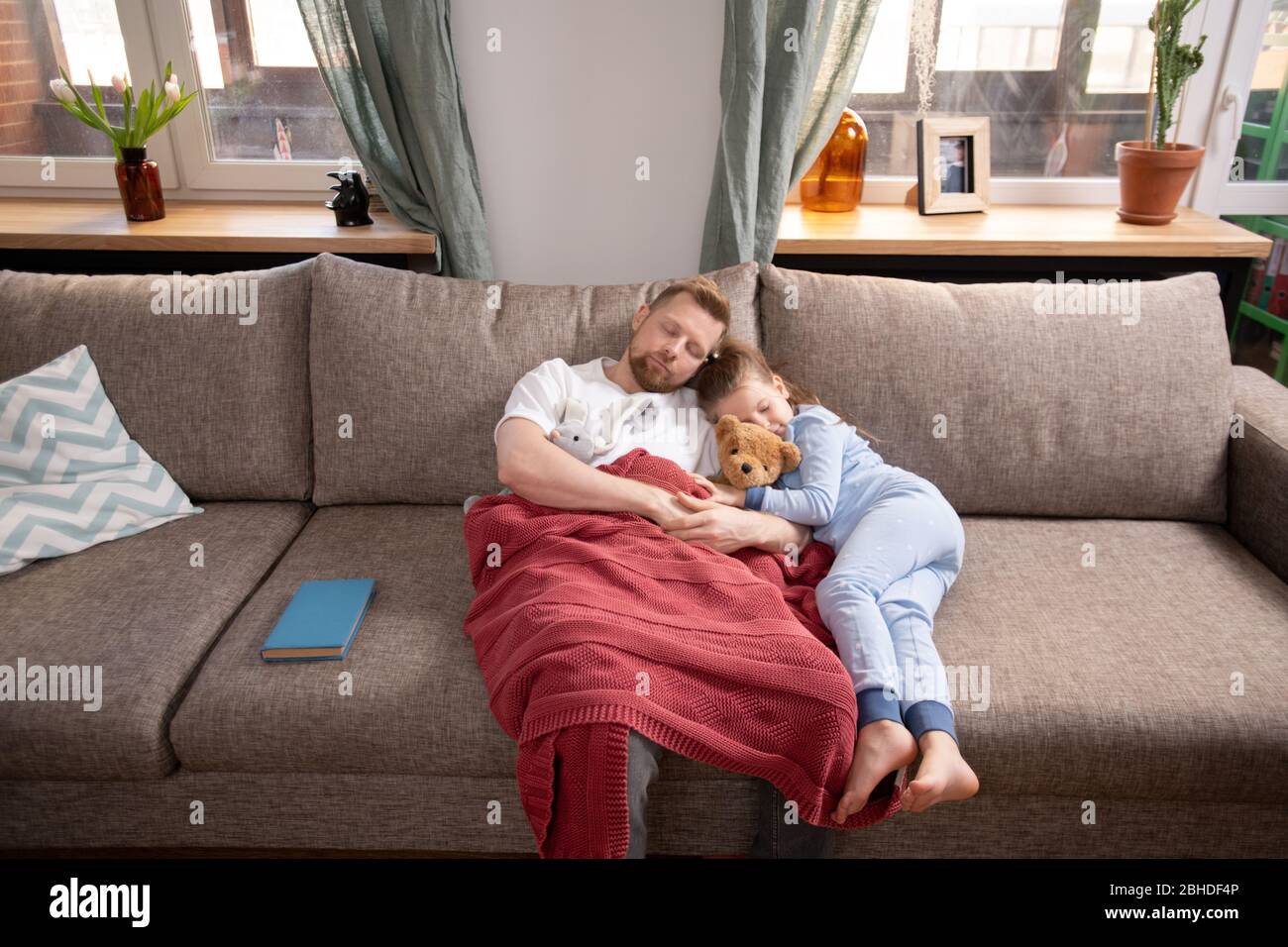 Müde junge Mann und seine süße kleine Tochter in blauen Pyjama liegen auf einer weichen Couch durch Fenster im Wohnzimmer und Nickerchen nach dem Abendessen Stockfoto