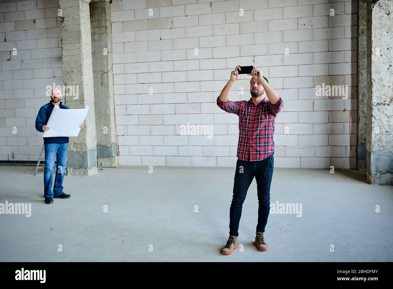 Junger Unternehmer mit Smartphone, der das unfertige Gebäudeinnere fotografiert, während sein Kollege im Hintergrund Blaupause hält Stockfoto