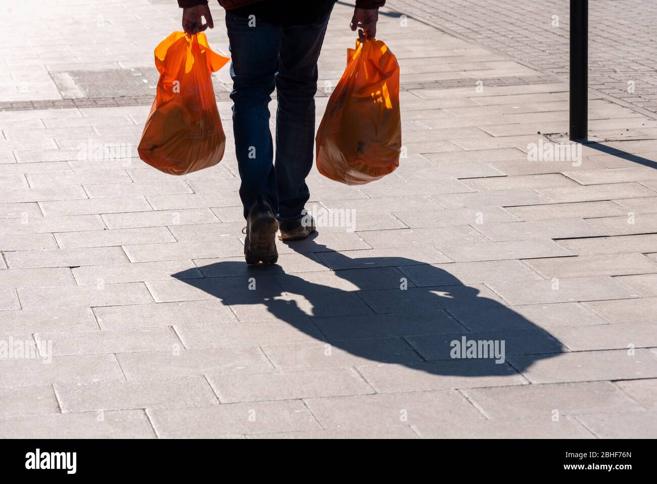 Shopper mit Plastiktüten am späten Nachmittag mit langem Schatten zu Fuß. Heller sonniger Tag. Orangefarbene Tragetasche. COVID-19 Sperrzeit. Schatten Stockfoto