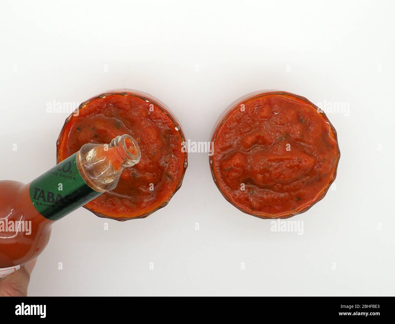 Draufsicht auf zwei Schalen mit frischem Tomatensalsa, eine mit Tabasco-Sauce, die in ihn gegossen wird, auf EINEM weißen Hintergrund. Stockfoto