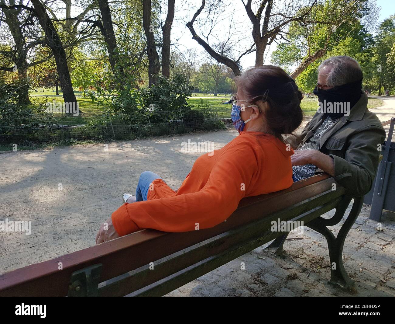 Altes Paar / Großeltern ( Mann / Mann & Frau / Frau ) mit Gesichtsmaske ( OP-Maske ), auf Parkbank sitzend - Covid-19 Corona Virus Lockdown Stockfoto