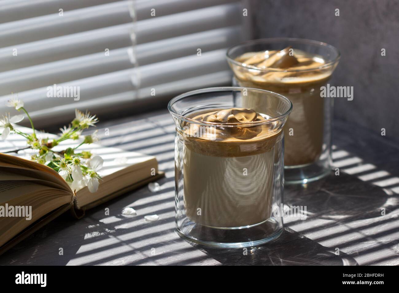 Dalgona Kaffee in transparenten Gläsern auf einer Fensterbank, die von Sonnenlicht beleuchtet wird. Stockfoto