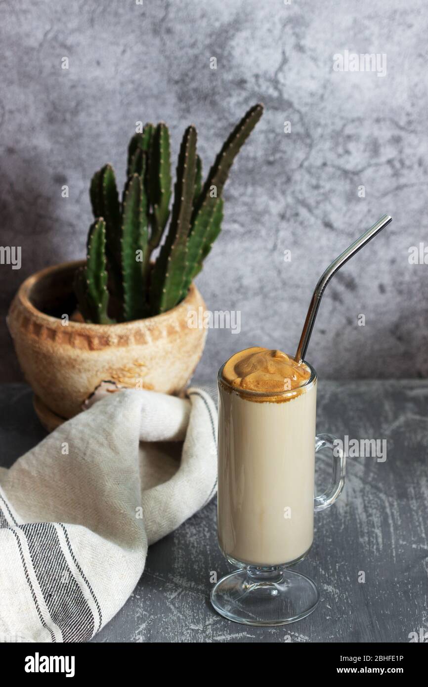 Dalgona Kaffee in einer transparenten Tasse mit einem Cocktail Stroh und Kaktus auf einem Betontisch. Stockfoto