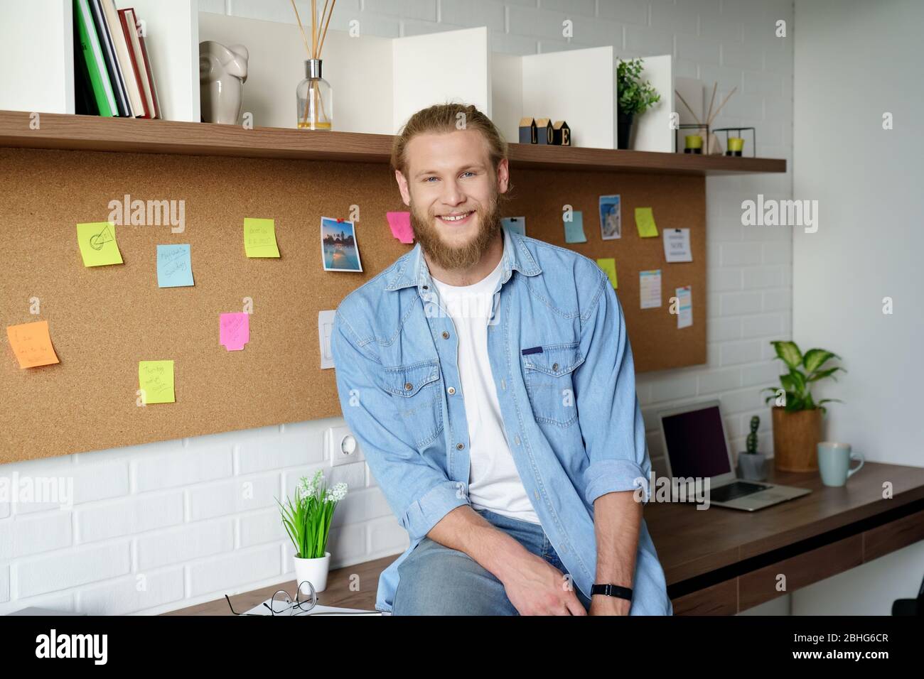 Lächelnder junger Erwachsener bärtiger Gelegenheitsmann, der Kamera im Heimbüro anschaut. Stockfoto