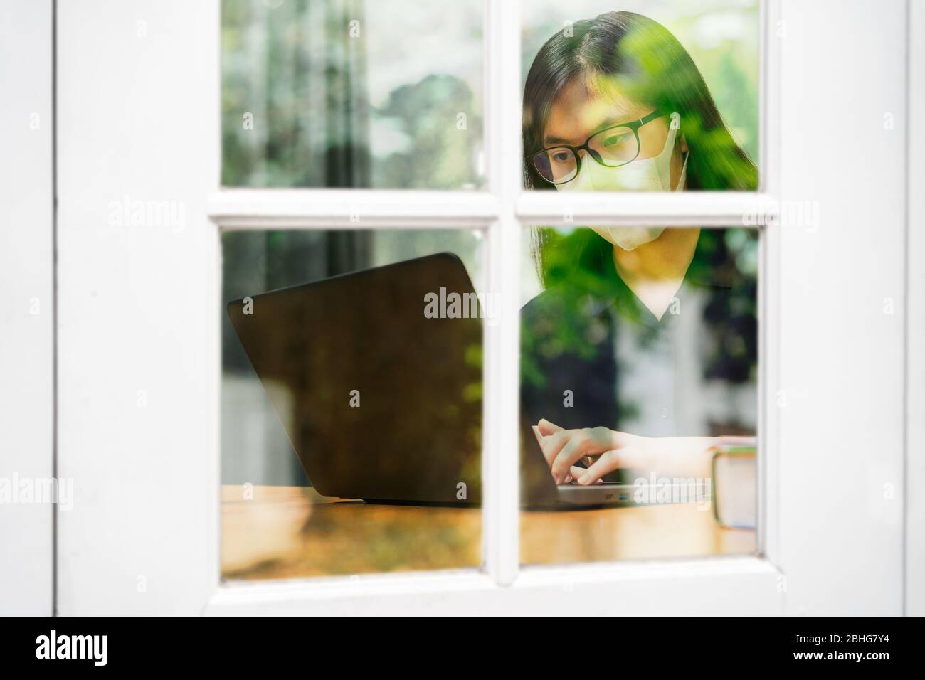 Junge Asiatin mit N95 Maske und Brille, die zu Hause an ihrem Computer arbeitet, durch Fenster mit Spiegelung von Bäumen gesehen, während der Stadtsperre Stockfoto