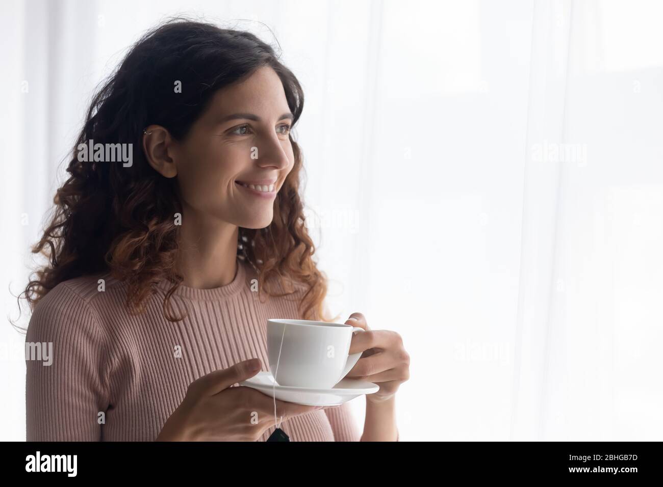 Glückliche junge hübsche Dame, die heißen Tee trinkt, beim Blick auf das Fenster. Stockfoto