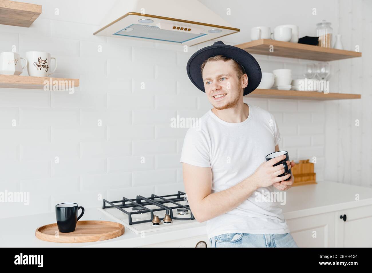 Junger Mann in Hut Lächeln trinken frischen Kaffee in Keramik schwarzen Becher. Helle Küche Hintergrund Stockfoto