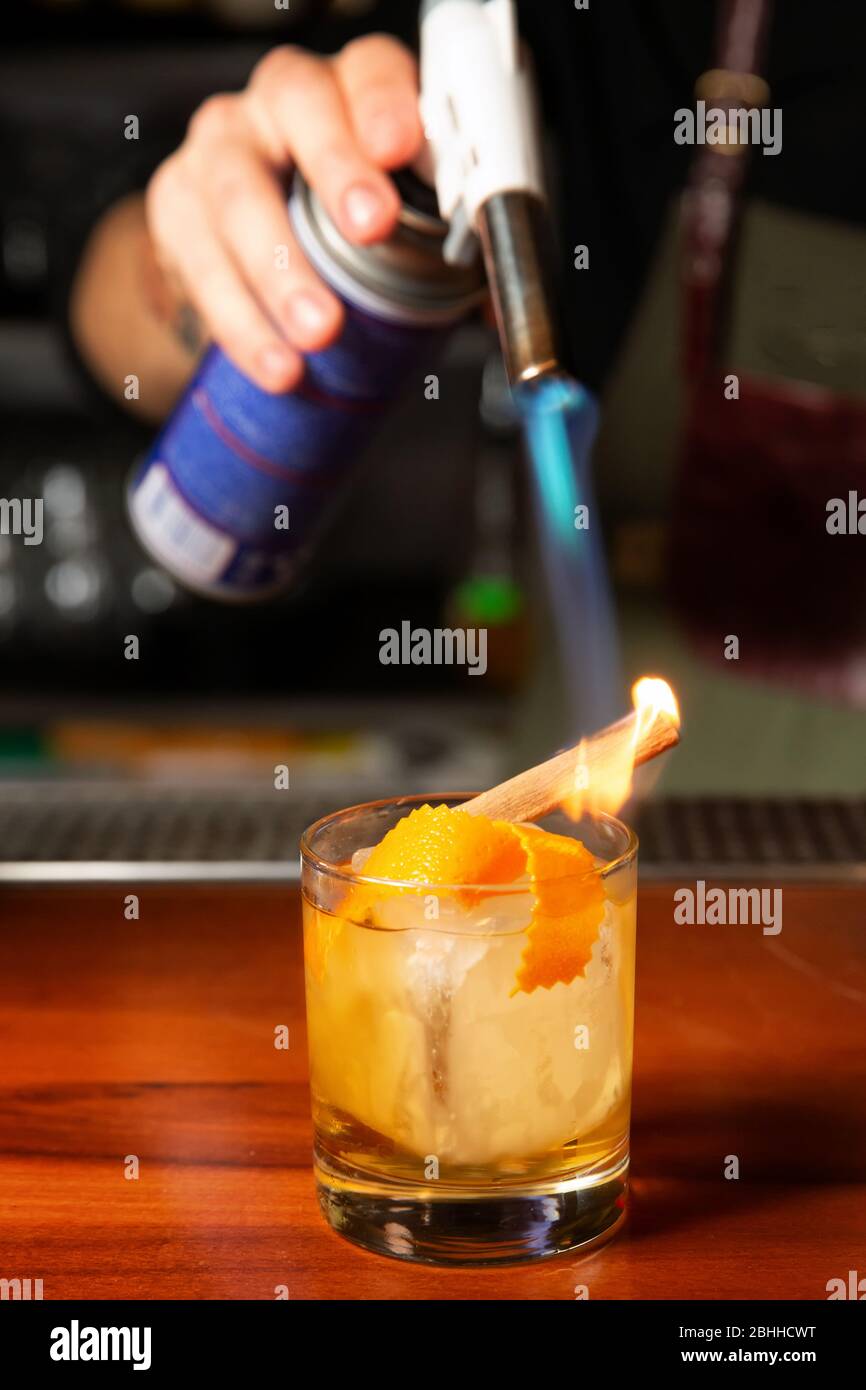 Ein männlicher Barkeeper verbrennt einen Zimtstab mit einem Gasbrenner in einer Bar. Prozess der Zubereitung eines Cocktails. Stockfoto