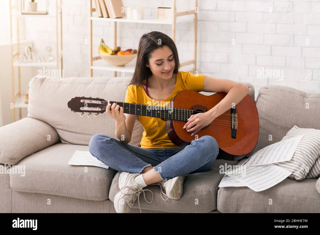 Bleiben Sie zu Hause, viel Spaß. Schöne musikalische Mädchen mit Noten spielen akustische Gitarre auf bequemen Sofa drinnen Stockfoto