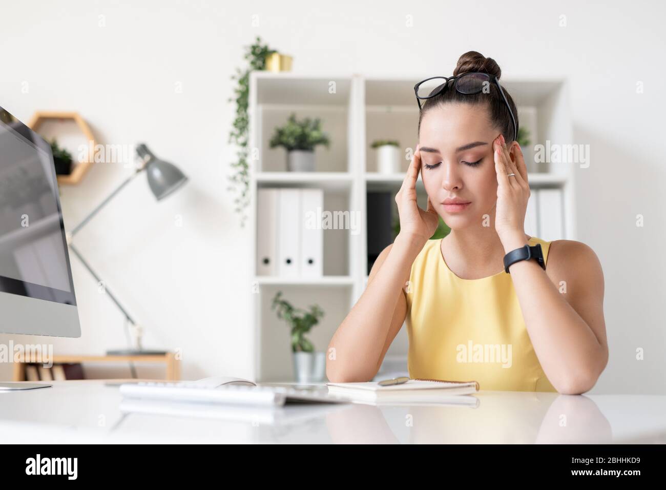 Müde junge Geschäftsfrau mit Brille auf dem Kopf sitzt am Schreibtisch im Büro und massiert Tempel, während Kopfschmerzen Stockfoto