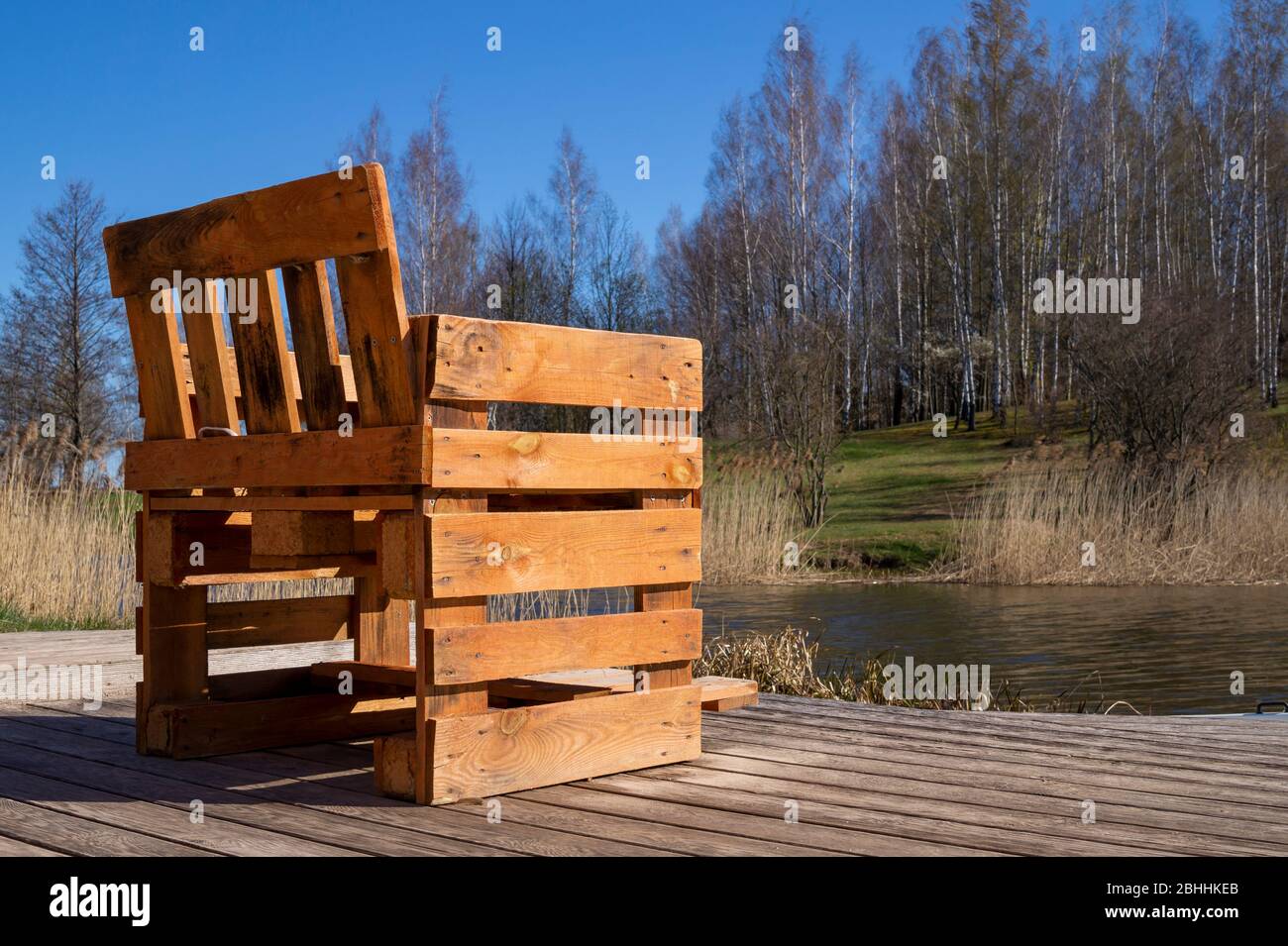 Rustikaler handgefertigter Stuhl aus Holzpaletten auf Holzdeck mit Blick auf einen Fluss oder See mit Schilf bei Frühlingssonne Stockfoto