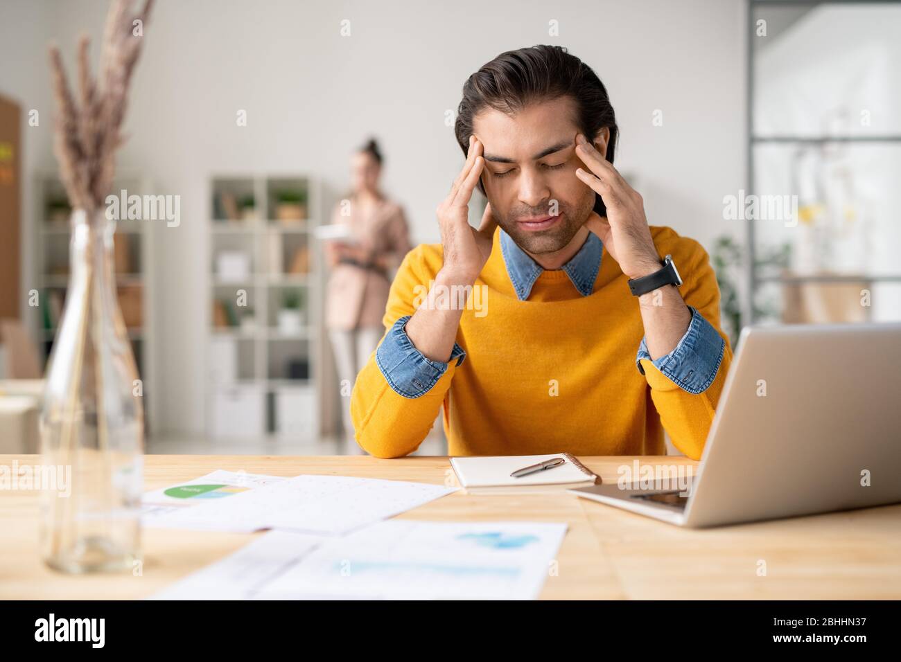 Verwirrt junger Mann in gelben Pullover sitzt am Schreibtisch mit Notizblock und hält den Kopf in den Händen, während Kopfschmerzen bei der Arbeit leiden Stockfoto