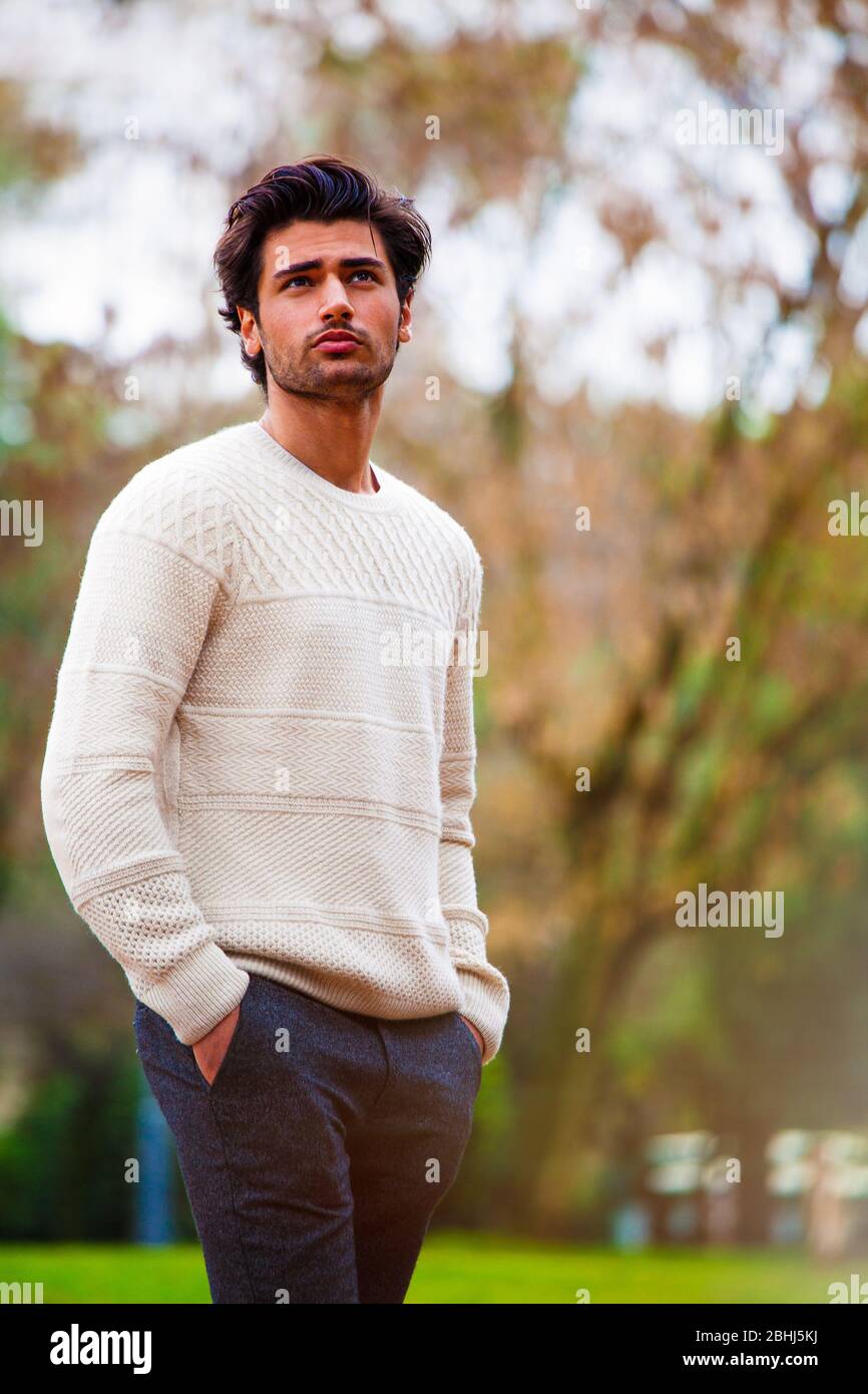 Schöner junger Mann draußen auf der Straße mit Händen in den Taschen. Mode Frisur, selbstbewusste Haltung. Weißer Pullover. Stoppeln. Stockfoto