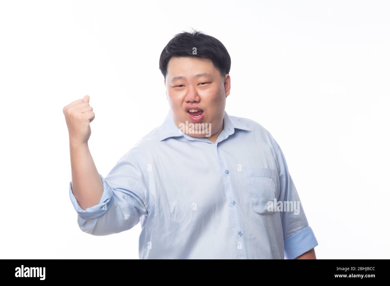 Aufgeregt asiatischen Fat man in blauem Hemd heben seine Faust mit glücklich erfreut Gesicht, feiern Erfolg isoliert auf weißem Hintergrund. Stockfoto