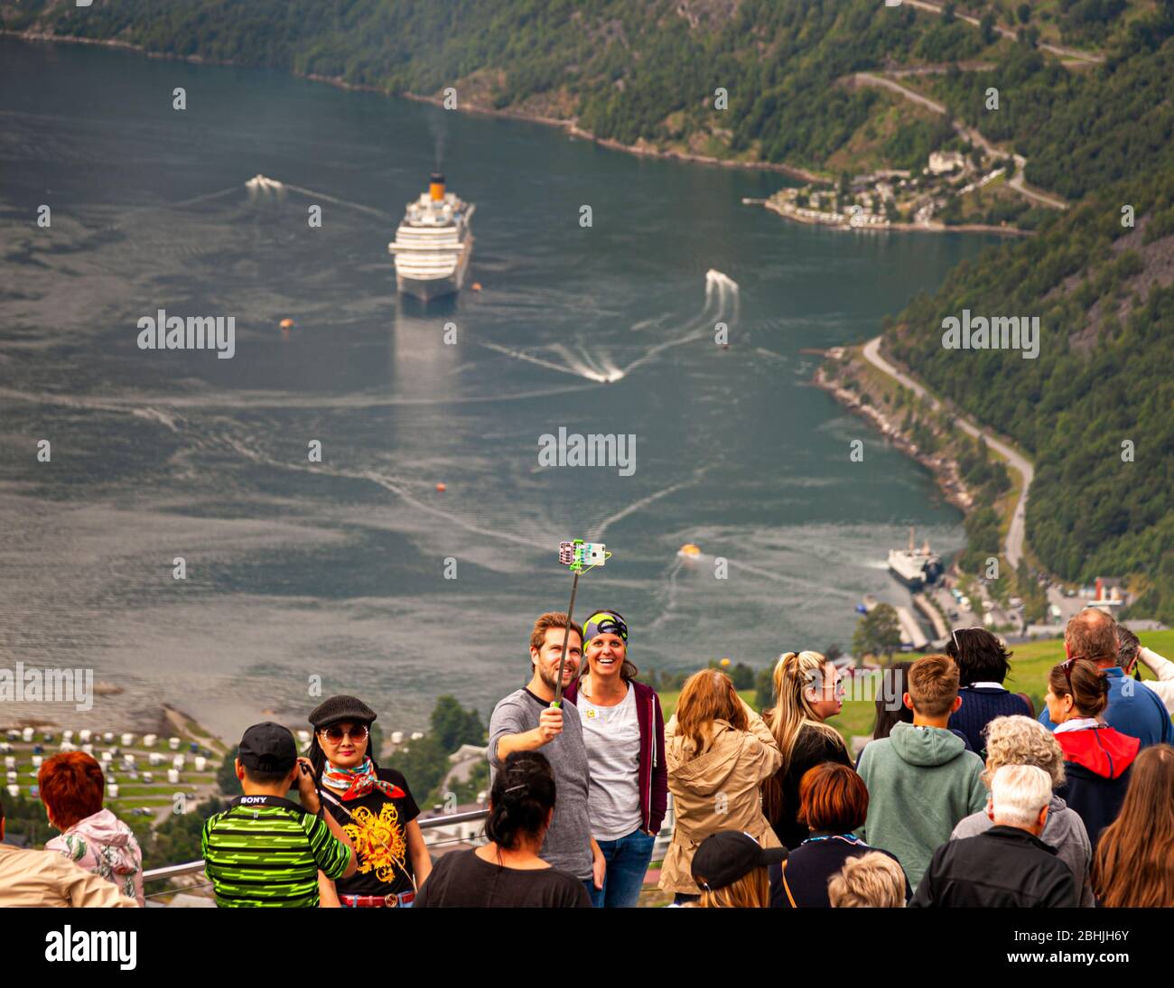 Am Aussichtspunkt Ørnesvingen über Geiranger, Norwegen, macht ein Paar ein Selfie mit einem Selfiestick inmitten einer dichten Menschenmenge von Besuchern. Unten liegt ein Kreuzfahrtschiff im Fjord, wobei ein kleineres Schiff zusätzlich am Kai festgemacht ist. Stockfoto