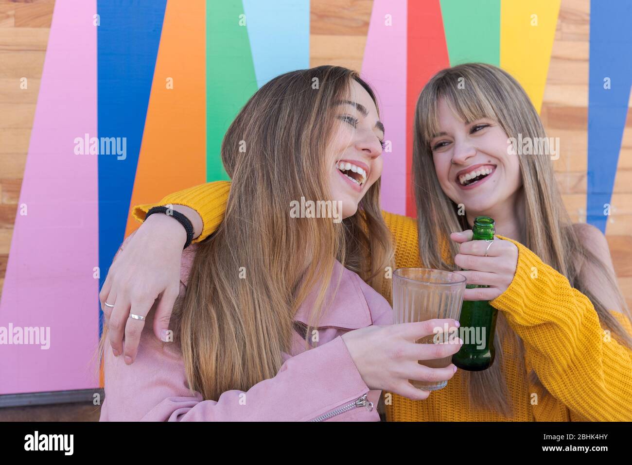 Zwei fröhliche junge kaukasische Frauen stoßen mit einem Drink an die bunte Wand einer Stadtbar Stockfoto