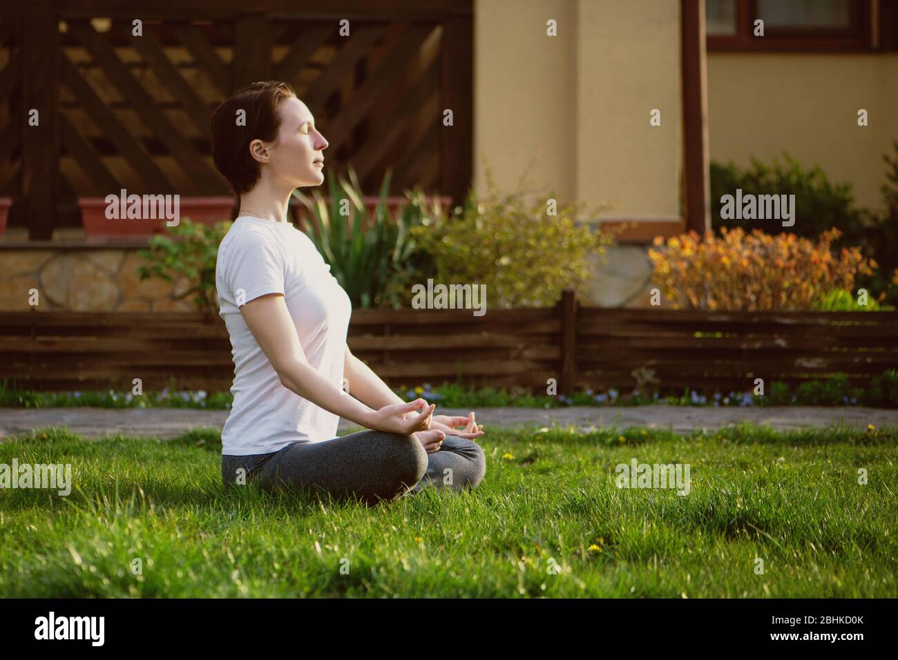 Eine junge Frau meditiert auf Gras im Hof neben dem Haus. Stockfoto