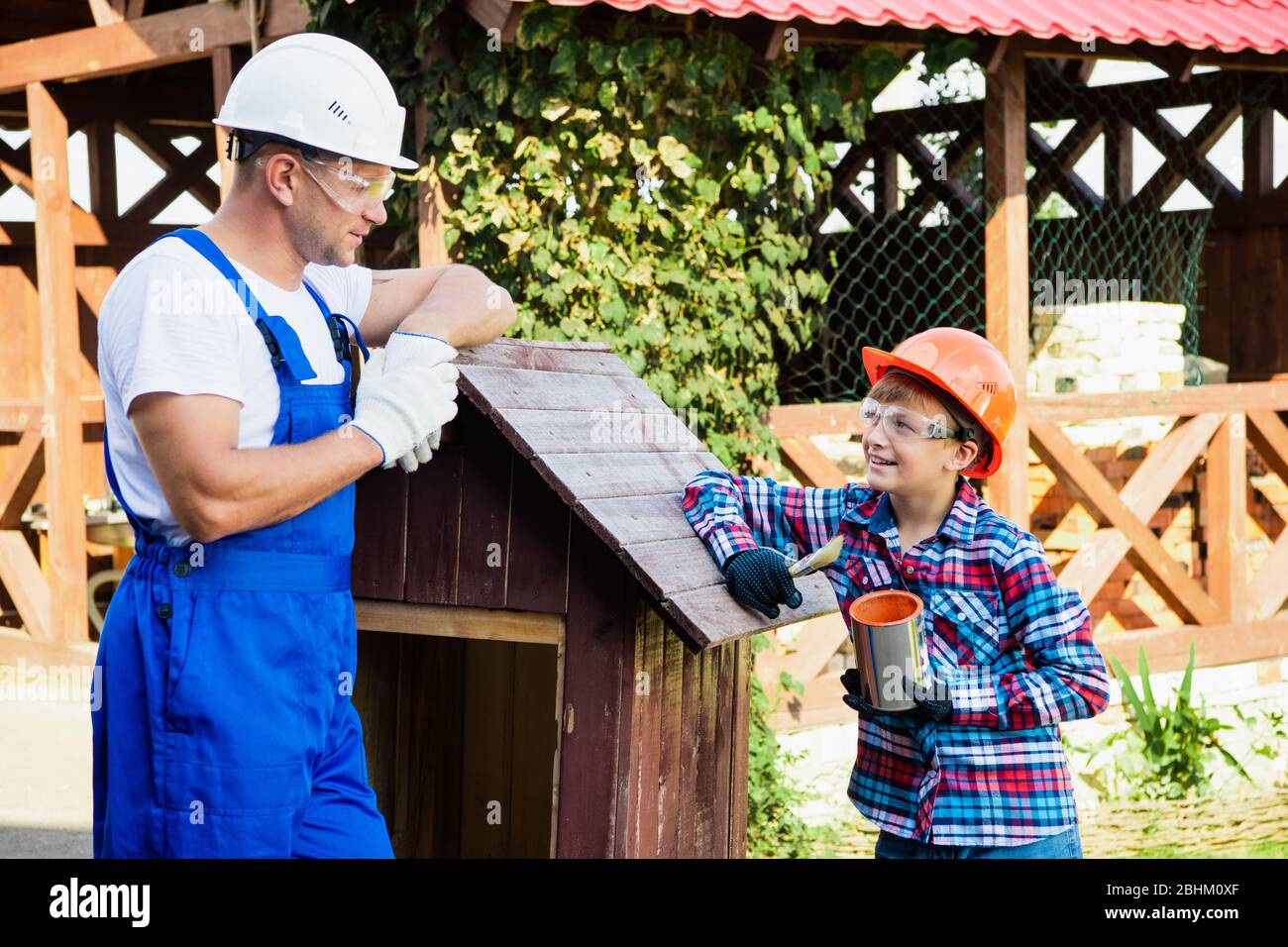 Vater und Sohn arbeiten zusammen in einer Holzwerkstatt. Sie konzentrieren sich auf den Bau eines hölzernen Hundehauses, der Sohn sitzt darin mit einer Farbe Stockfoto